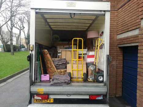 The image shows the open rear of a white delivery truck parked on a paved driveway adjacent to a brick building, with a patch of grass and trees visible in the background. Inside the truck, various types of rubbish are temporarily stored, including upholstered furniture such as a patterned armchair and a fabric-covered cushion, along with cardboard boxes, a pink plastic container, and a small portable fan or heater. There are also yellow hand trucks, a small black and red trolley, and a portable vacuum cleaner positioned amid the clutter. The truck's interior ceiling is plain, with a hook hanging down from the top. The overall scene reflects a typical rubbish removal scenario, associated with commercial waste clearance services in the Crystal Palace area, highlighting the collection of mixed domestic and light commercial waste material scheduled for disposal or recycling by Commercial Waste Crystal Palace.