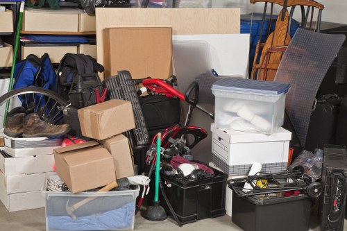 A cluttered indoor storage area filled with various items prepared for rubbish removal, including cardboard boxes, plastic storage containers, and black and grey rubbish bins. The cardboard boxes are stacked and vary in size, some sealed with tape. The clear plastic storage container contains miscellaneous objects, while the larger boxes and bins hold assorted household waste. A bicycle wheel is partially visible on the left side of the image, with other tools and items scattered amongst the collection. The background includes shelving units and furniture, indicating a domestic or commercial storage setting, possibly in Crystal Palace. The lighting appears natural and even, highlighting the mixture of materials such as cardboard, plastic, and metal. The scene reflects an organized approach to waste collection, consistent with rubbish removal services provided by Commercial Waste Crystal Palace within the local postcode area.
