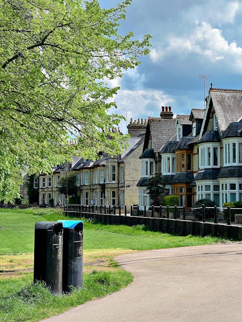 A wide view of a residential street showing a row of traditional Victorian-style terraced houses with bay windows, of varying shades of grey, beige, and brown brickwork, under a partly cloudy sky. In 