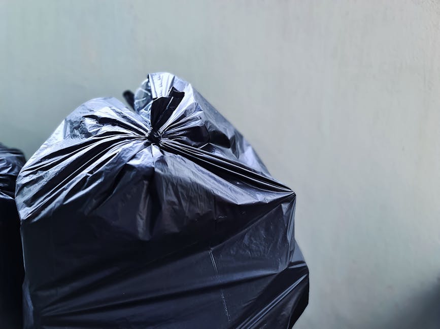 A close-up view of multiple black plastic rubbish bags filled with waste, tightly tied at the top, resting against a plain, light-colored wall. The bags have a shiny, crinkled texture and are placed o