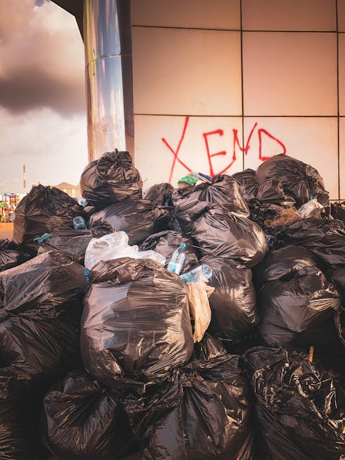A large pile of black plastic rubbish bags stacked against a modern building wall with tiled surface, some of which are torn open revealing plastic bottles and discarded packaging inside. The bags are